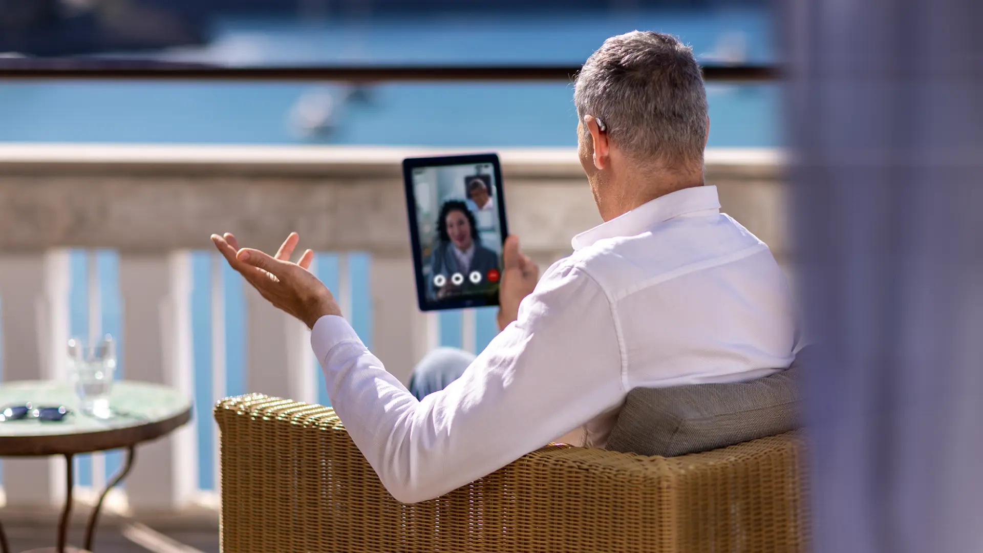 Homem com aparelho auditivo conversando por videochamada em um tablet, na varanda de casa